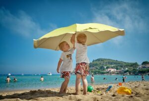Zwei Kinder stehen unter einem gelben Sonnenschirm am Strand.